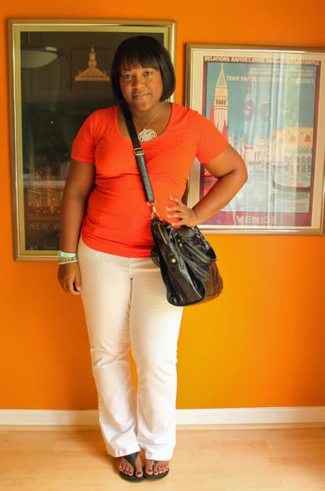 A black haired woman wearing an orange shirt and white pants standing and posing with her black bag with two portraits on the orange background
