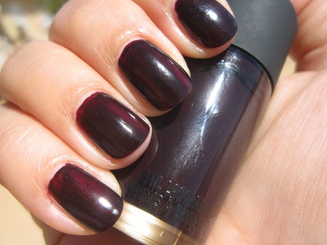 A hand with a dark purple creme nail polish holding a nail lacquer of the same shade