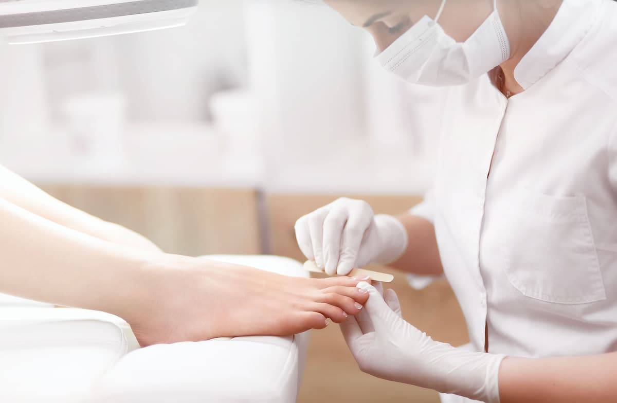 A masked woman filing another's toe nails during a medical pedicure
