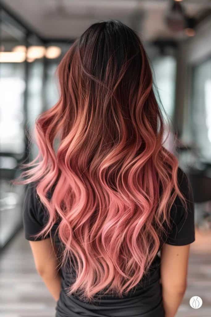 Woman with long wavy hair that transitions from brunette at the roots to rose gold towards the ends, standing indoors.