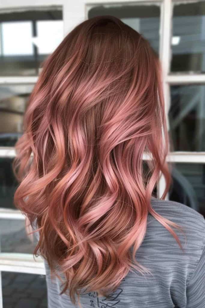 A woman with wavy, rose gold hair, viewed from the back, standing in front of a window with grid patterns.