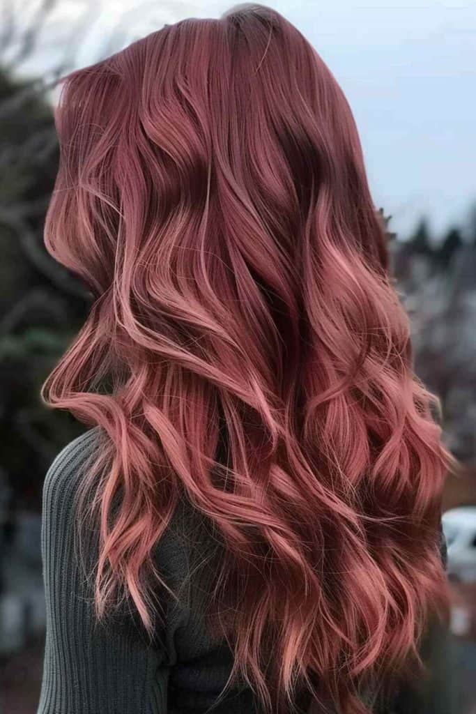 A woman with long, wavy rose gold hair, viewed from behind, standing outdoors with a blurred natural backdrop.