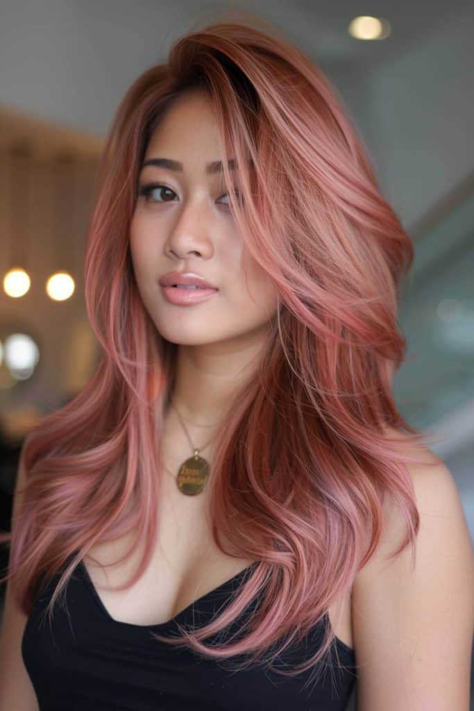 A woman with wavy, rose gold hair and a black top, looking at the camera with a subtle expression in a softly lit indoor setting.