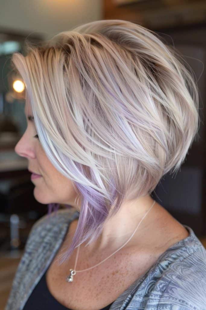 Close-up side profile of a person with a layered, asymmetrical, short stacked bob haircut featuring blonde and light purple highlights, wearing a gray top and a necklace.