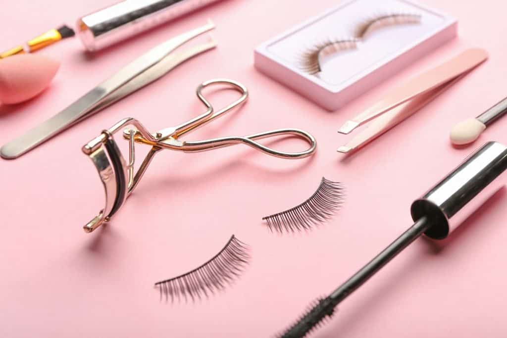 eyelash curler, tweezers and false lashes on a pink backdrop
