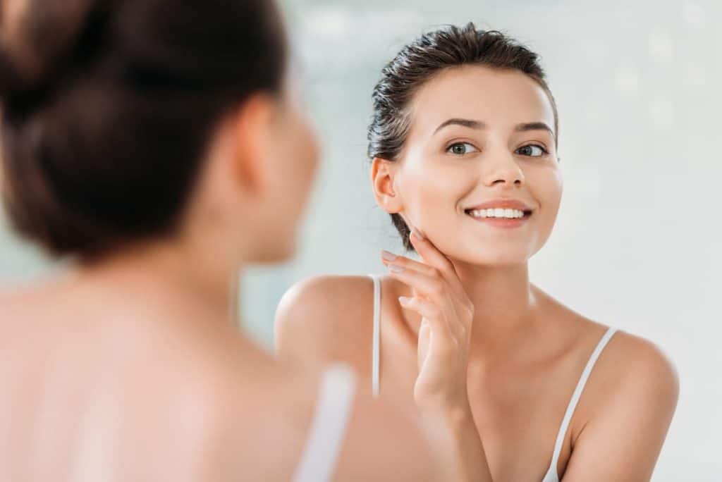 Beautiful smiling young woman touching skin and looking at mirror in bathroom