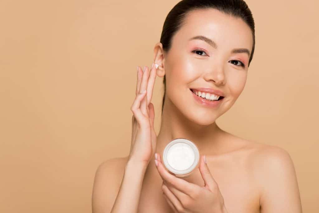 Asian woman holding a white face cream jar and applying cream to her face
