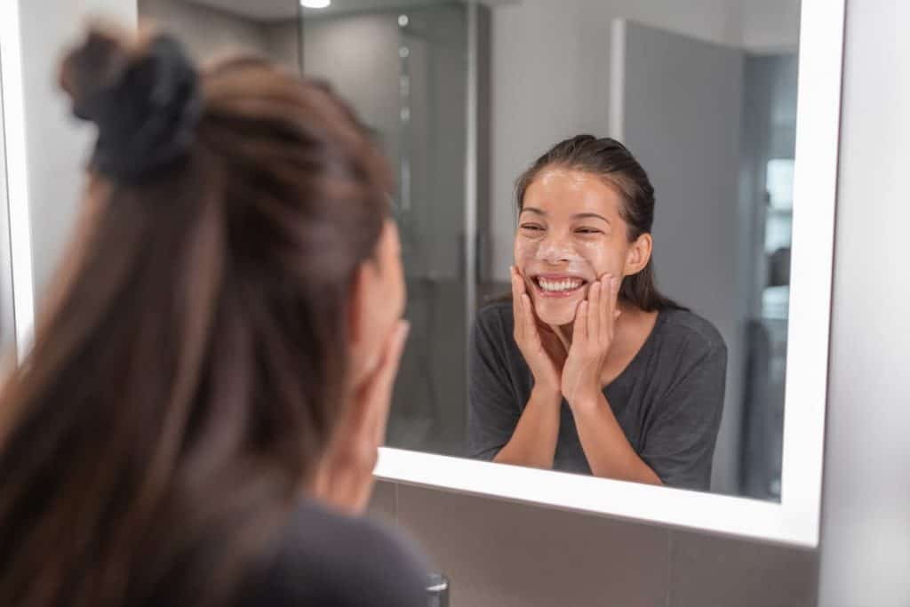 asian woman applying a facial scrub exfoliator while looking in the mirror