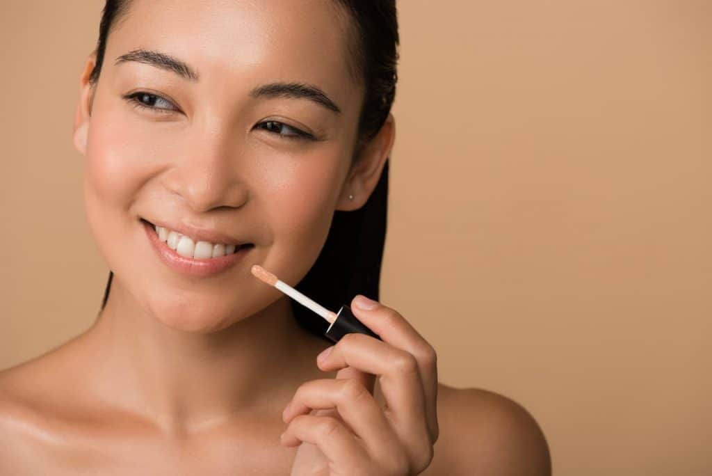 smiling Asian woman applying lip tint in a shade of beige
