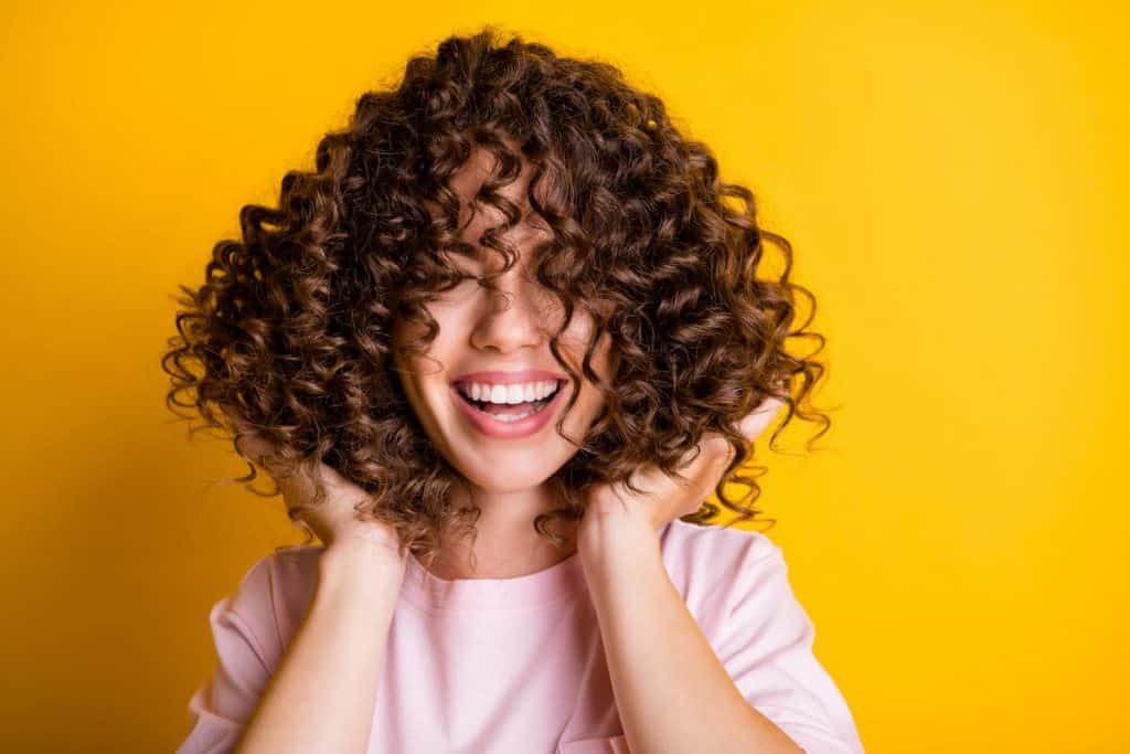 curly-haired smiling woman wearing pink shirt in a yellow background