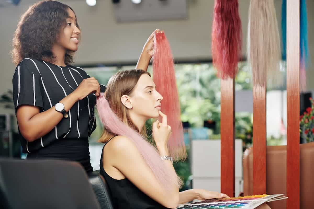 pensive young beautiful woman sitting in chair and looking at different hair extensions shown by hairstylist
