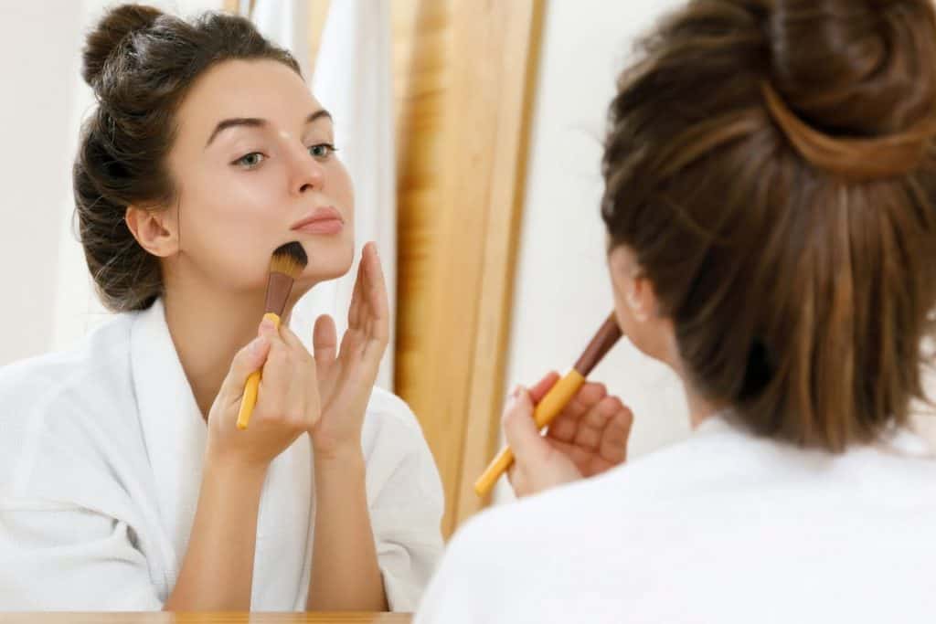 woman looking in the mirror and applying foundation on her face