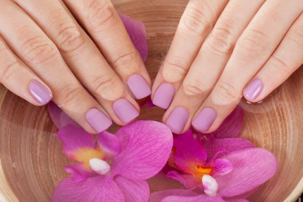 violet acrylic nails of female in a bowl of water with violet orchid flowers