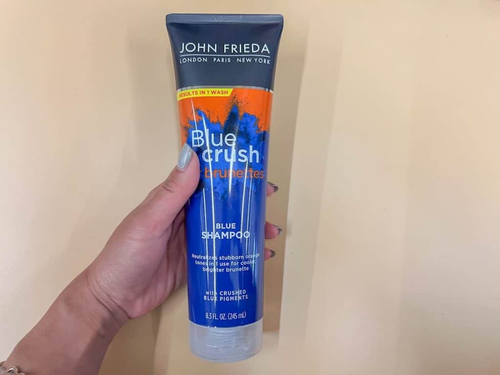 a woman's hand holding john frieda blue crush shampoo on a beige background