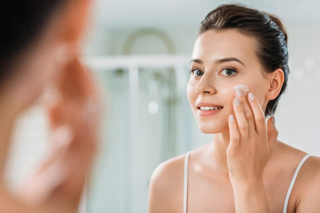 selective focus of smiling young woman applying face cream and looking at mirror