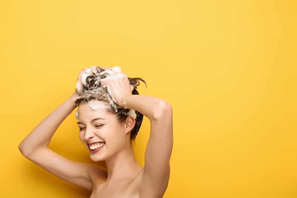 cheerful girl with closed eyes washing hair on yellow background
