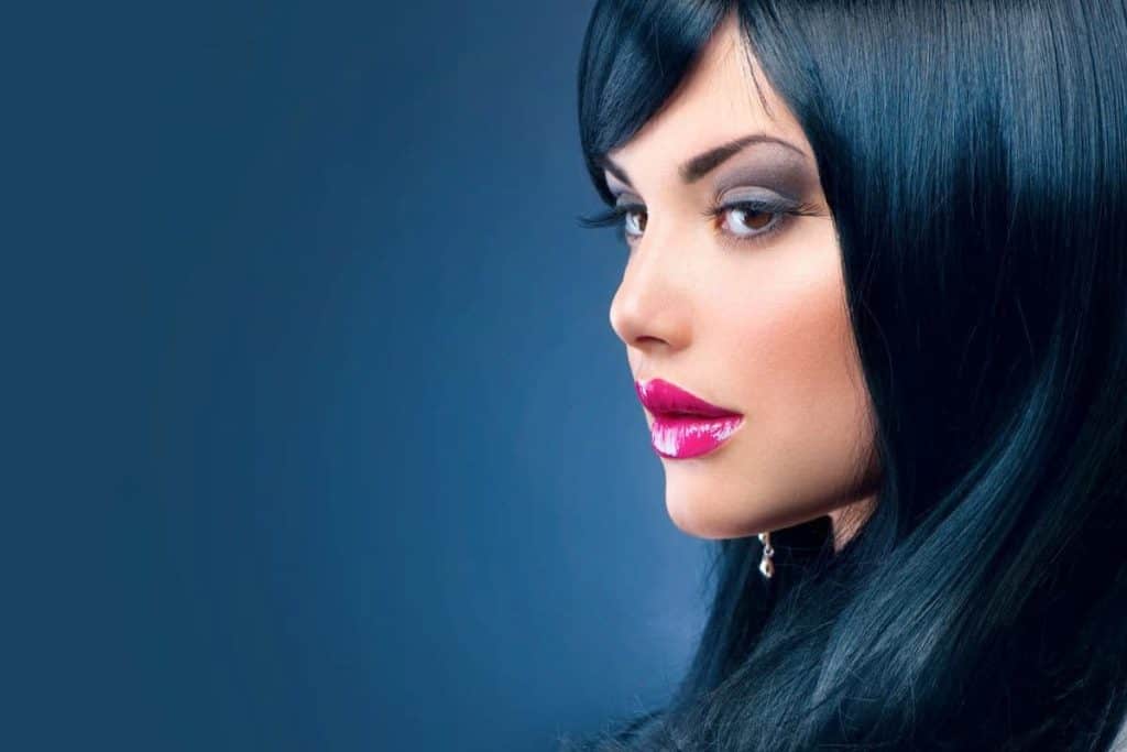 a sophisticated woman wearing black off shoulder and pink glossy lipstick looking sideways with long blue black hair