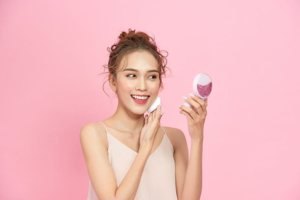 Asian woman holding a makeup brush on pink background.