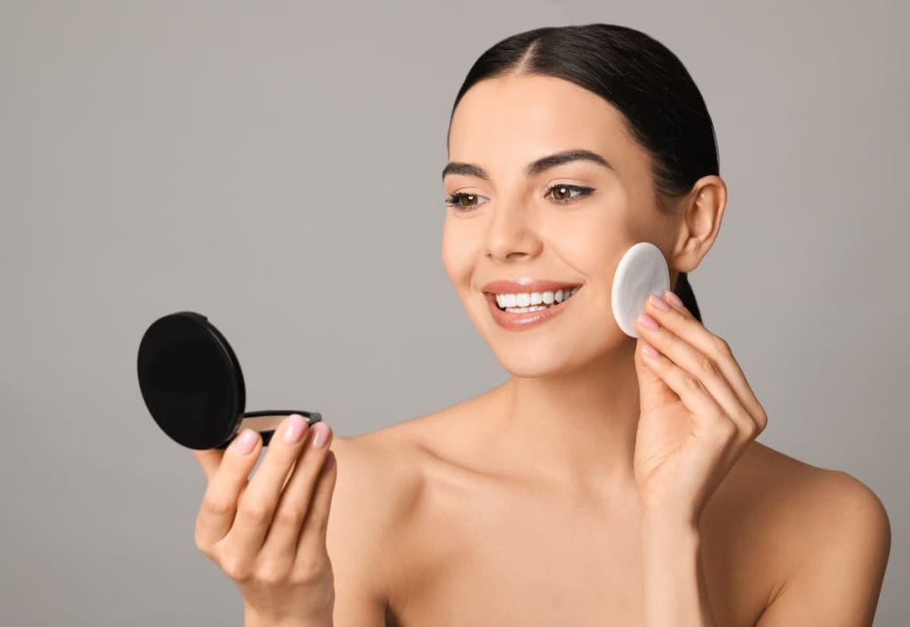 A young woman is applying pressed powder with a compact mirror.