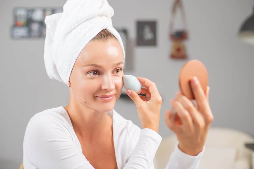 pretty woman with towel on head applying foundation or cream on face with a blotting sponge