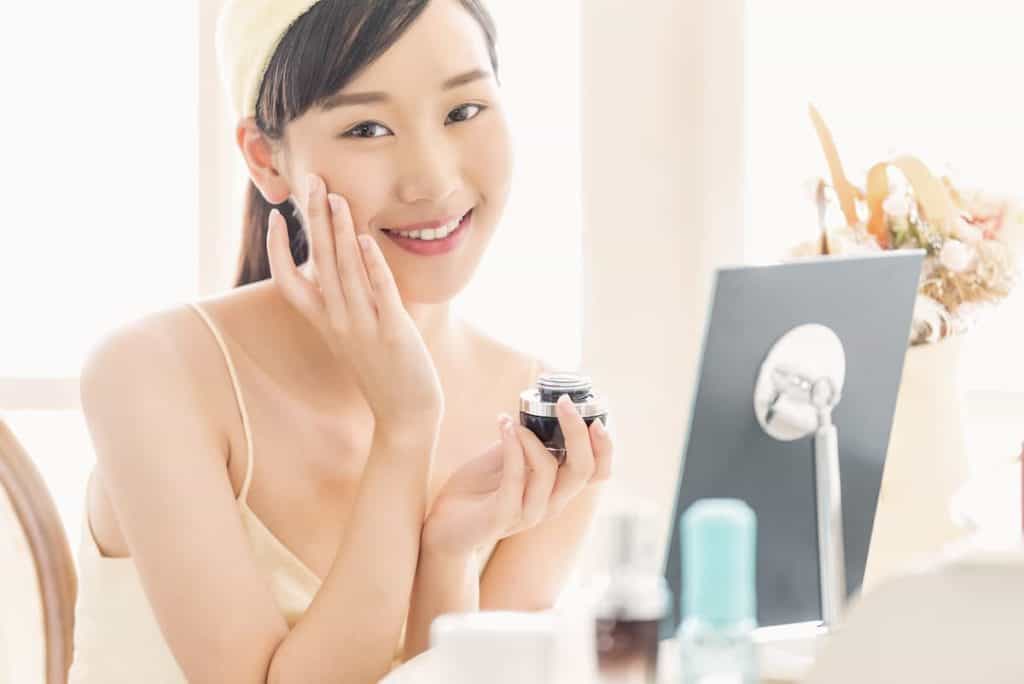 smiling young beautiful Asian woman with fresh healthy skin applying moisturizer