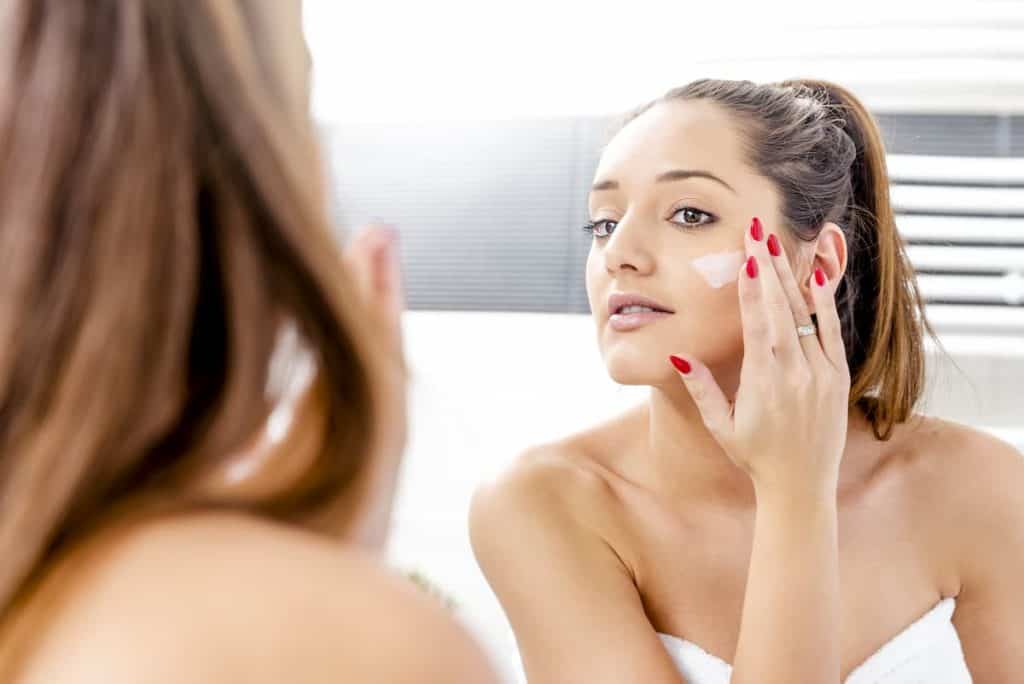 a woman in white towel applying azelaic acid on face in front of mirror