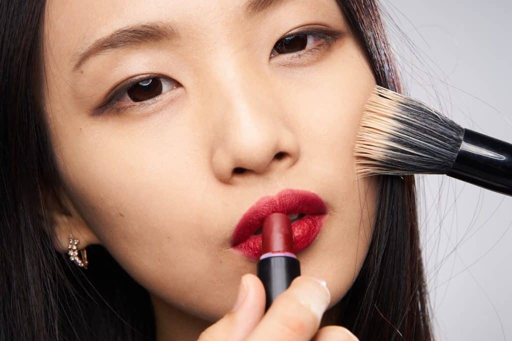 closeup view of an Asian woman applying red lipstick and foundation with a brush