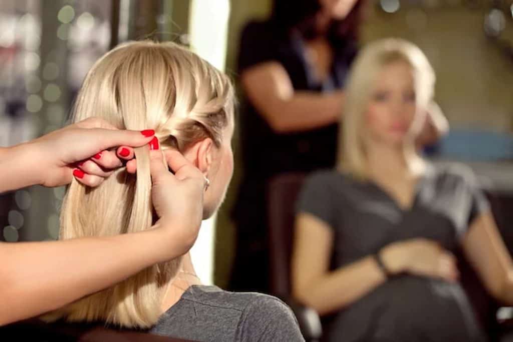 A blonde woman is sitting in front of a mirror while her hair is being braided
