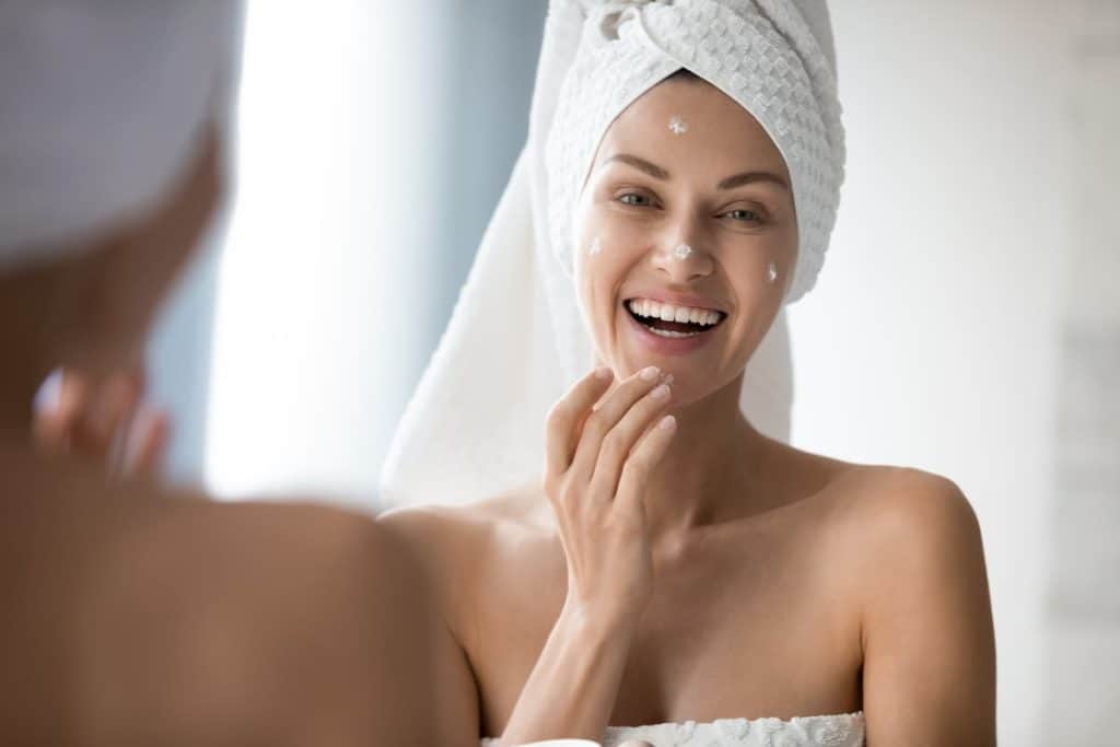 a woman with hair wrapped in white towel is laughing while applying moisturizer on face