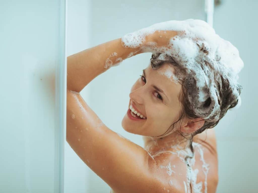 A laughing woman is washing her hair with shampoo in the shower.