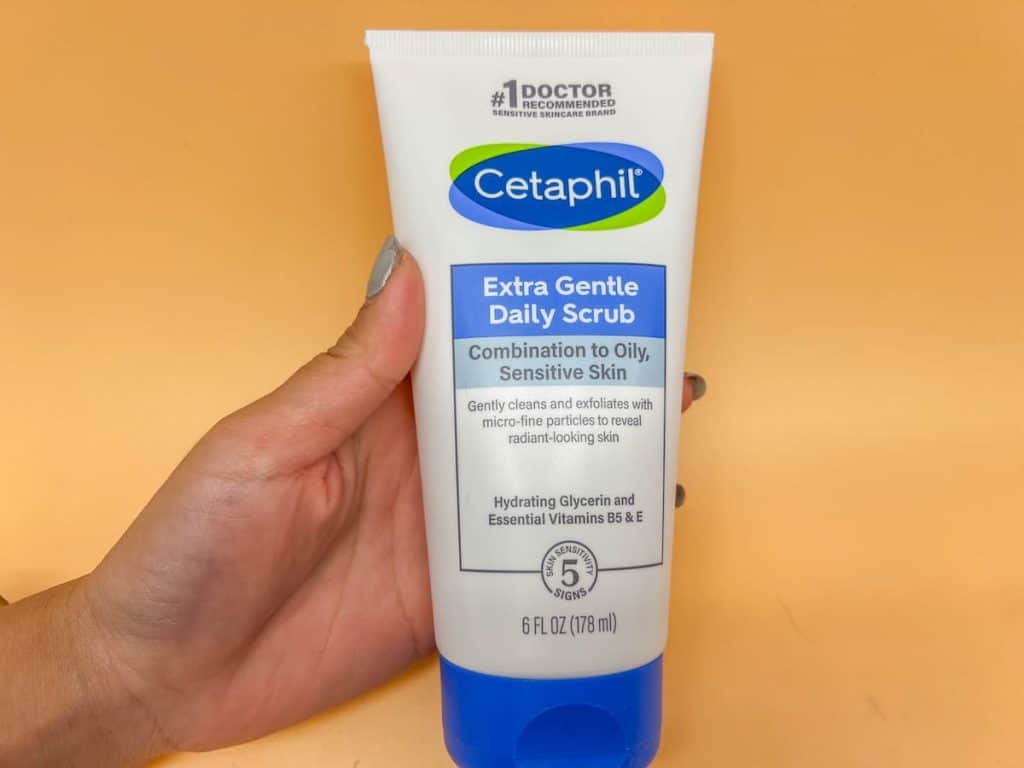 A woman's hand holding a cetaphil extra gentle daily scrub on a peach background