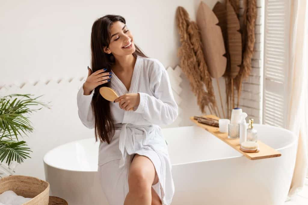 A woman wearing bathrobe is sitting in a bathtub brushing her long dark hair while smiling