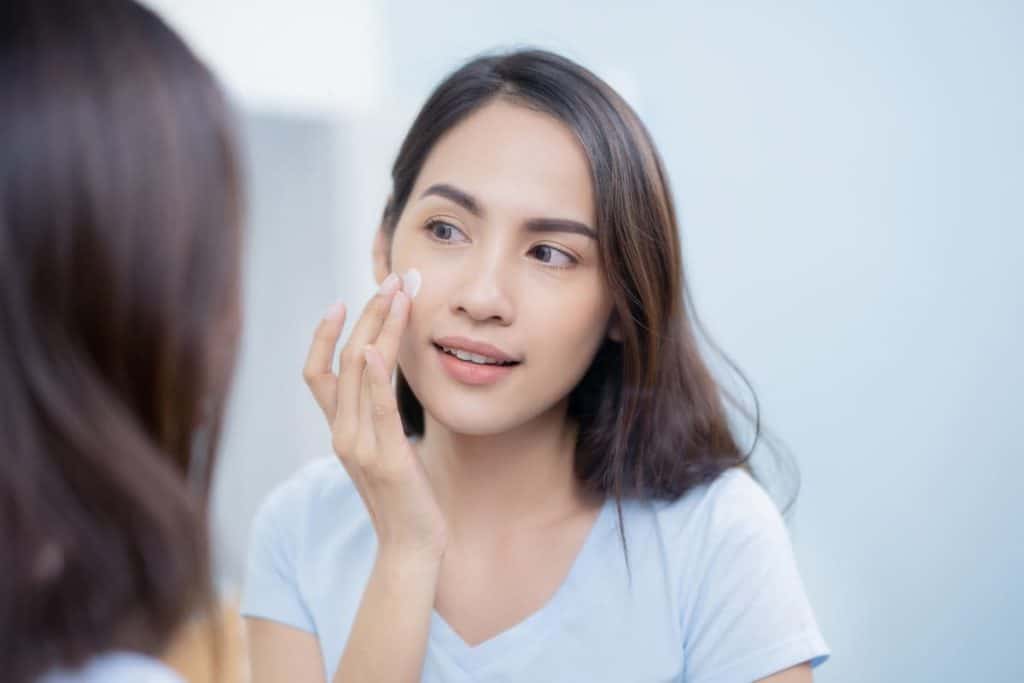 Asian woman applying cream to her face in the mirror.