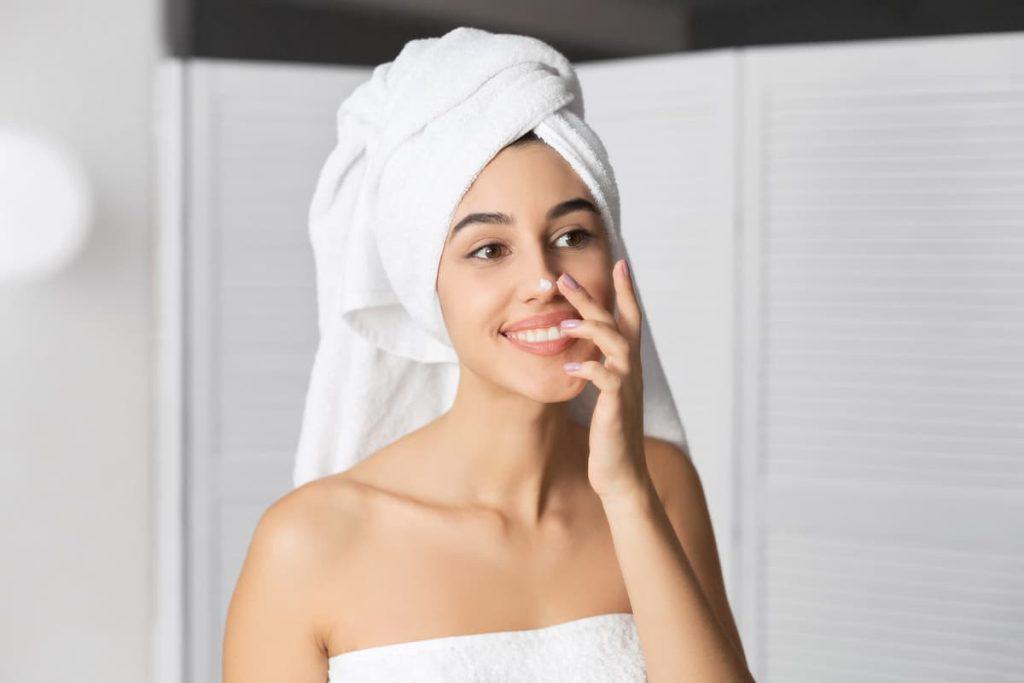 A young woman applying nose cream with a towel on her head is posing in front of a mirror.