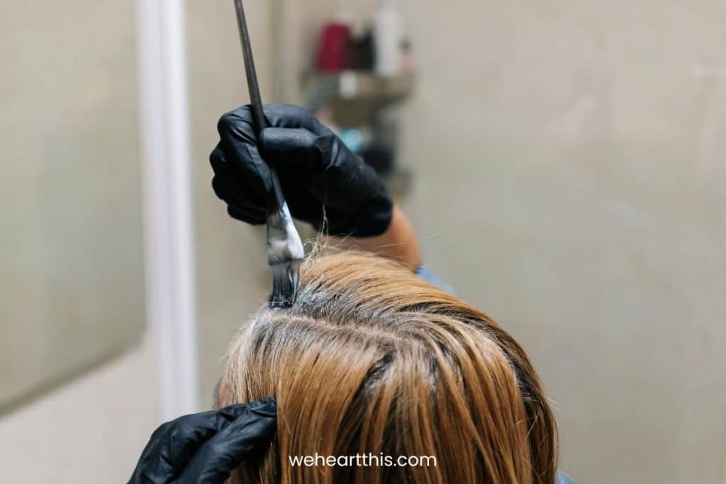 a woman applying box hair dye to her own hair using black brush at the bathroom 