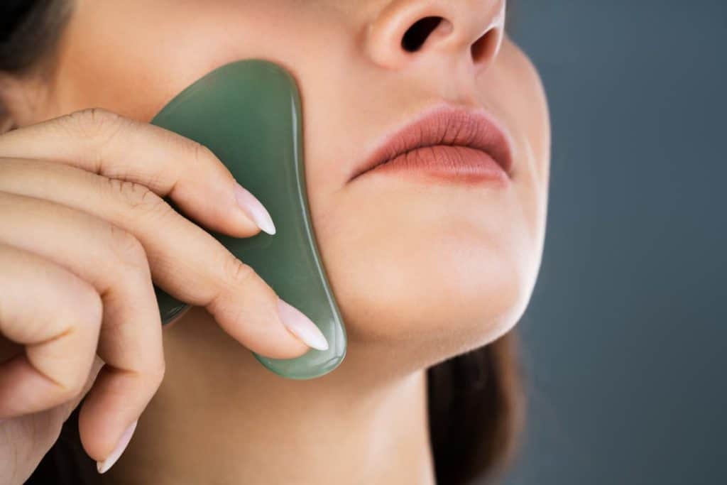 a close up image of a woman massaging her cheeks with gua sha after applying face oil