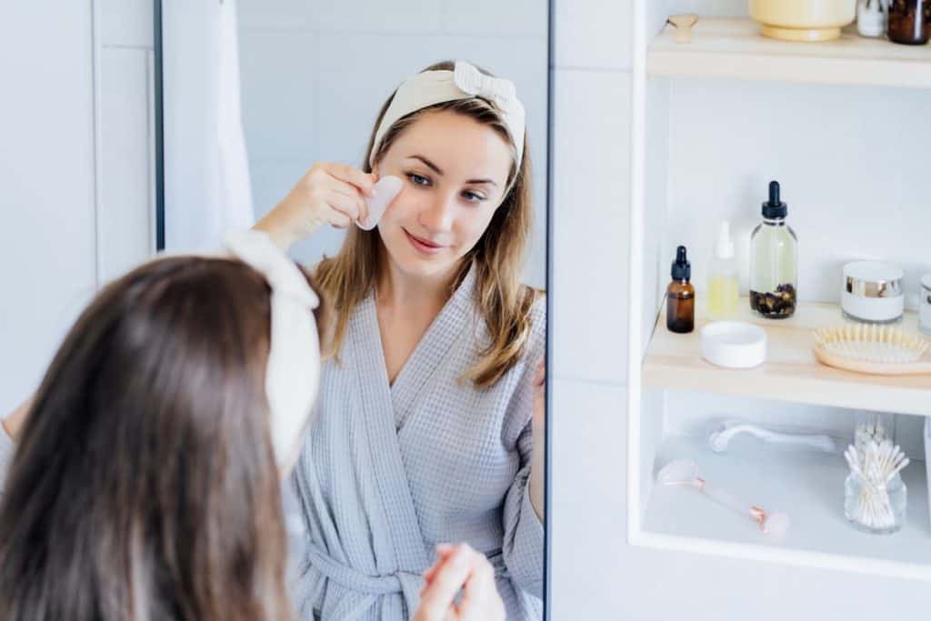 A woman in a bathrobe is using gua sha stone on her face in front of the bathroom with different oils on her side