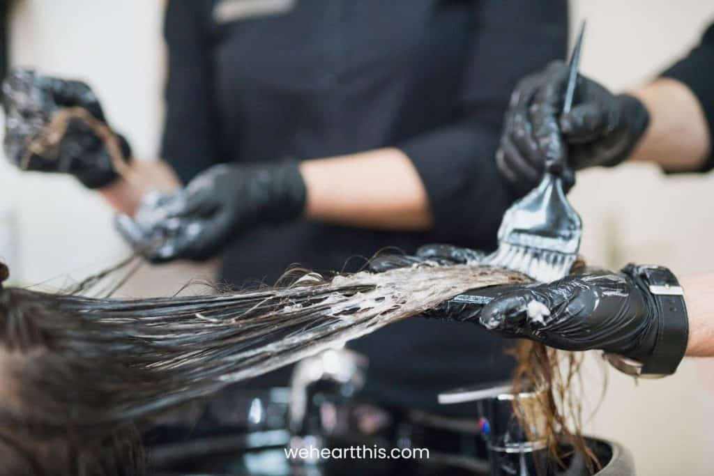 two hair stylist wearing black gloves are applying hair dye to their client using a black brush while wearing black apron
