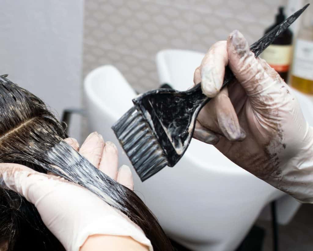 a hair stylist wearing black gloves are applying hair dye to their client using a black brush while wearing black apron