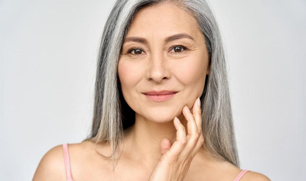 a beautiful old woman with long natural gray hair smiling at the camera while touching her cheeks