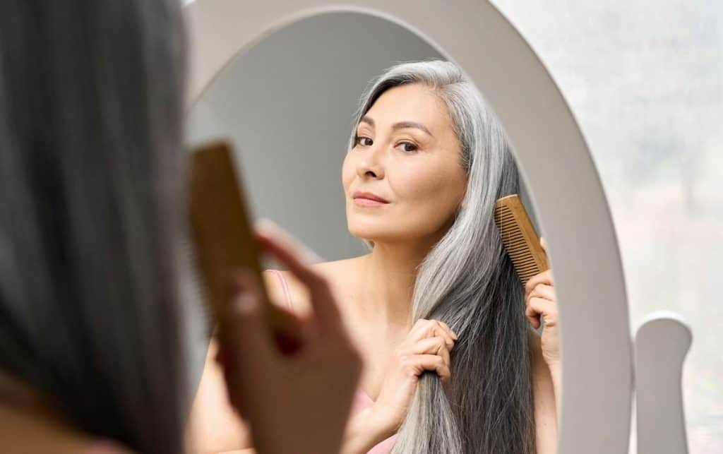 a middle-aged woman brushing her natural gray hair with wooden brush infront of a mirror