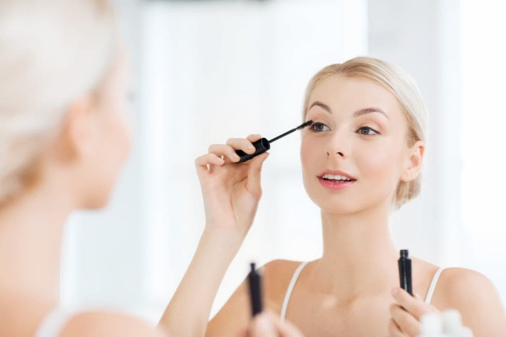 A blonde woman looking at the mirror while applying a mascara