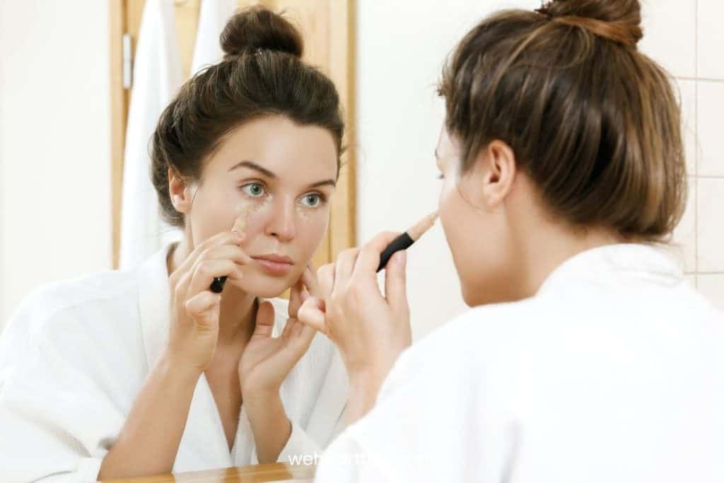 a woman wearing white bathrobe applying concealer under her eyes infront of bathroom mirror