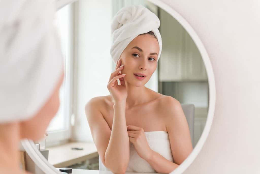 A woman in a towel is looking at herself in the mirror while touching her face