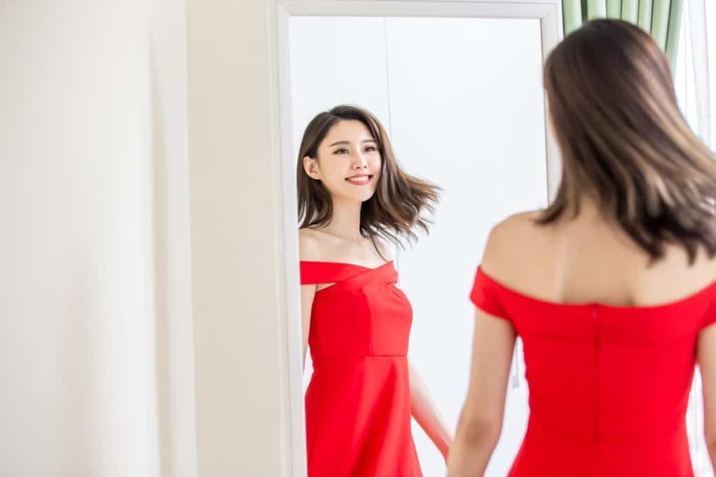 A woman in a red dress looking at herself in the mirror while smiling.