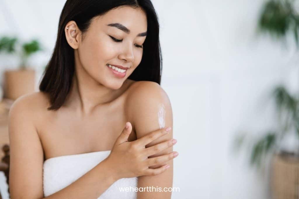 a young woman with short black hair and body wrapped in a white towel is applying inviscrepe body balm on her shoulder