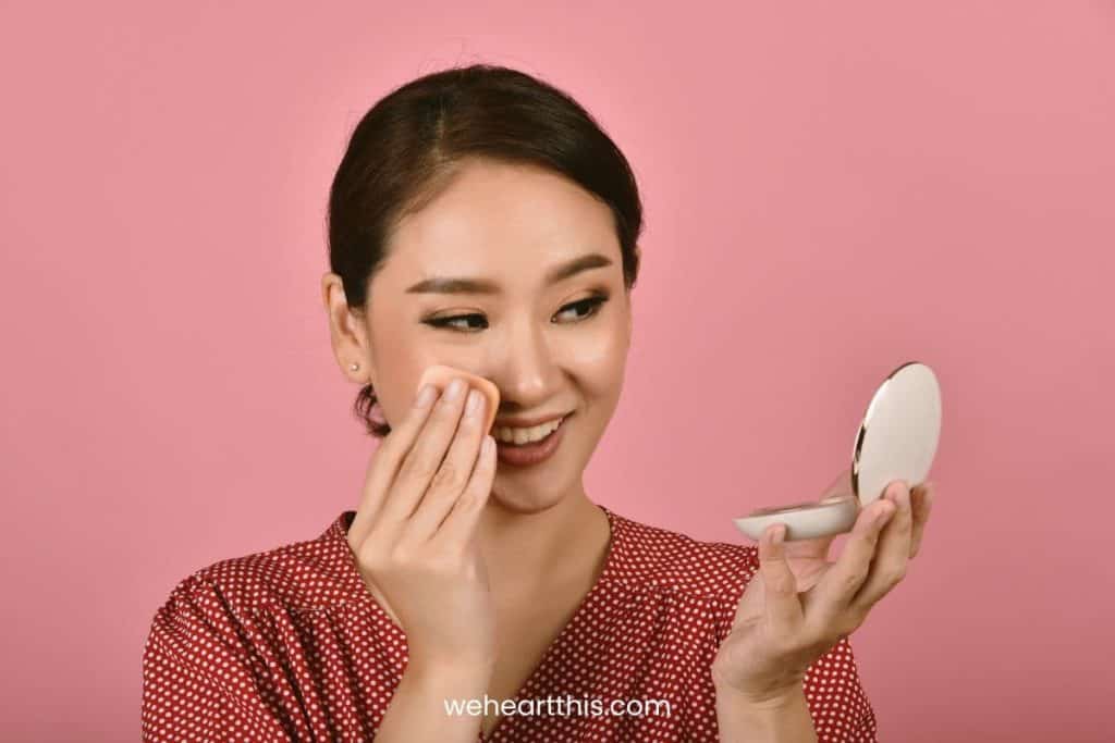 an asian woman wearing mustard polka dots top is applying pressed powder on her face happily