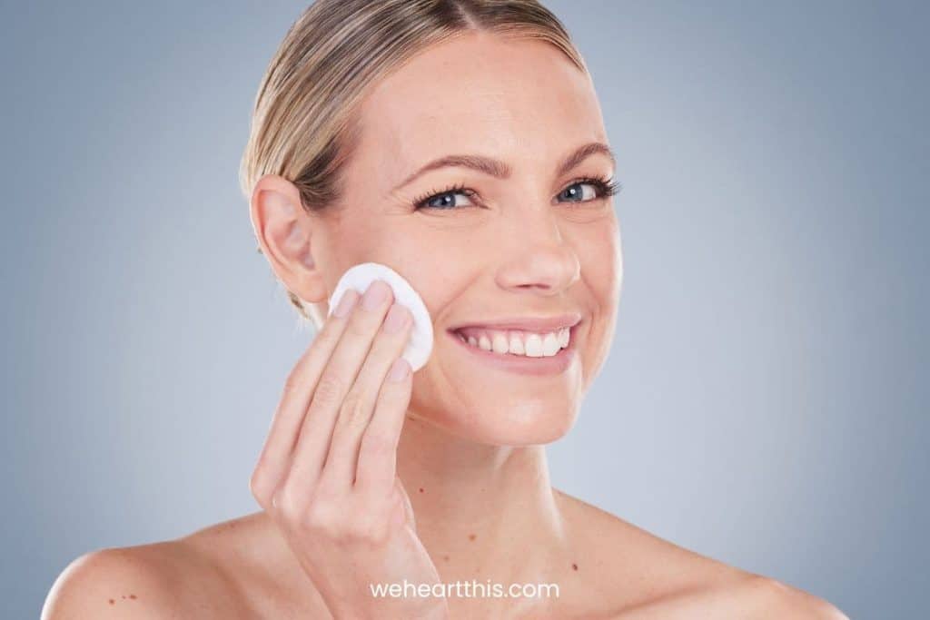 A woman smiling while using cotton to her face with toner in a dark background