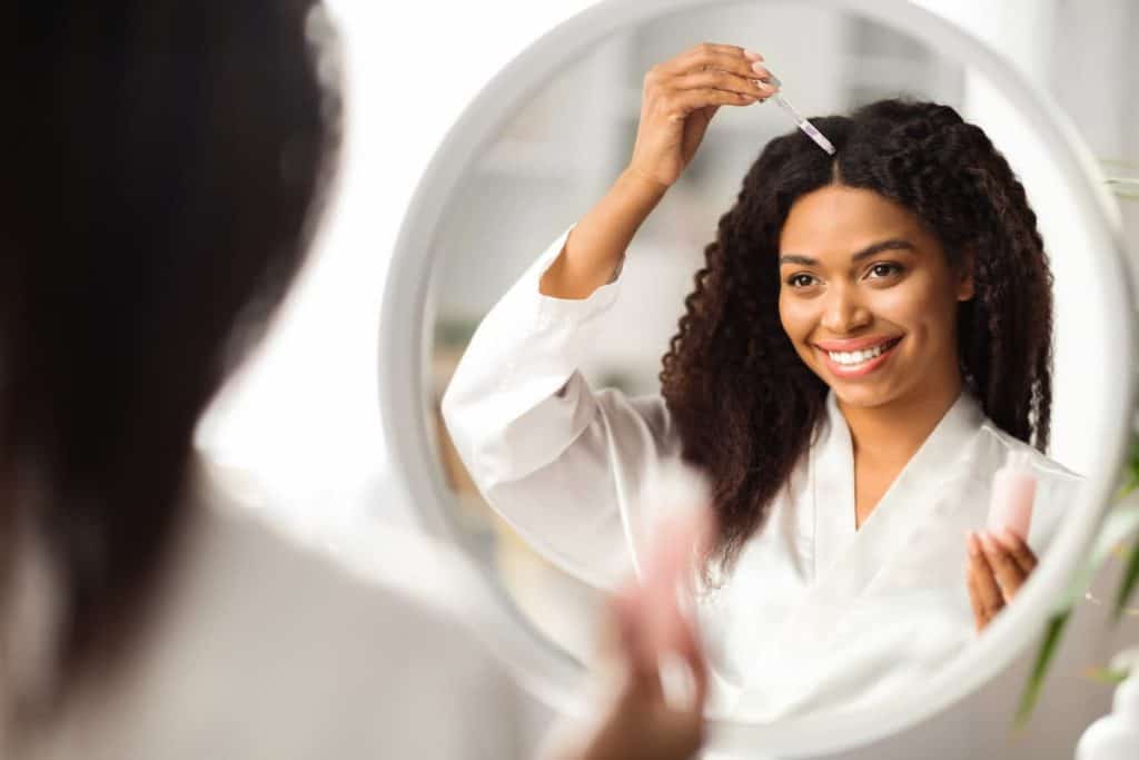 A smiling woman with curly hair is applying hair oil in front of a mirror.