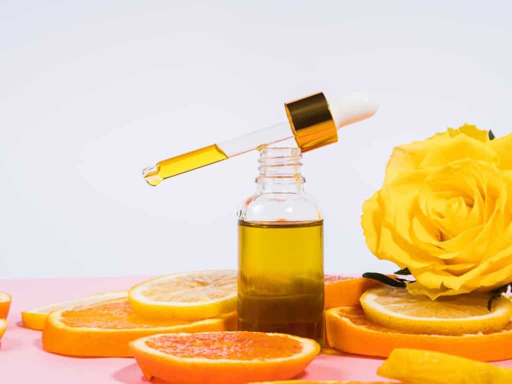 A bottle of vitamin c serum and orange slices on a pink background.